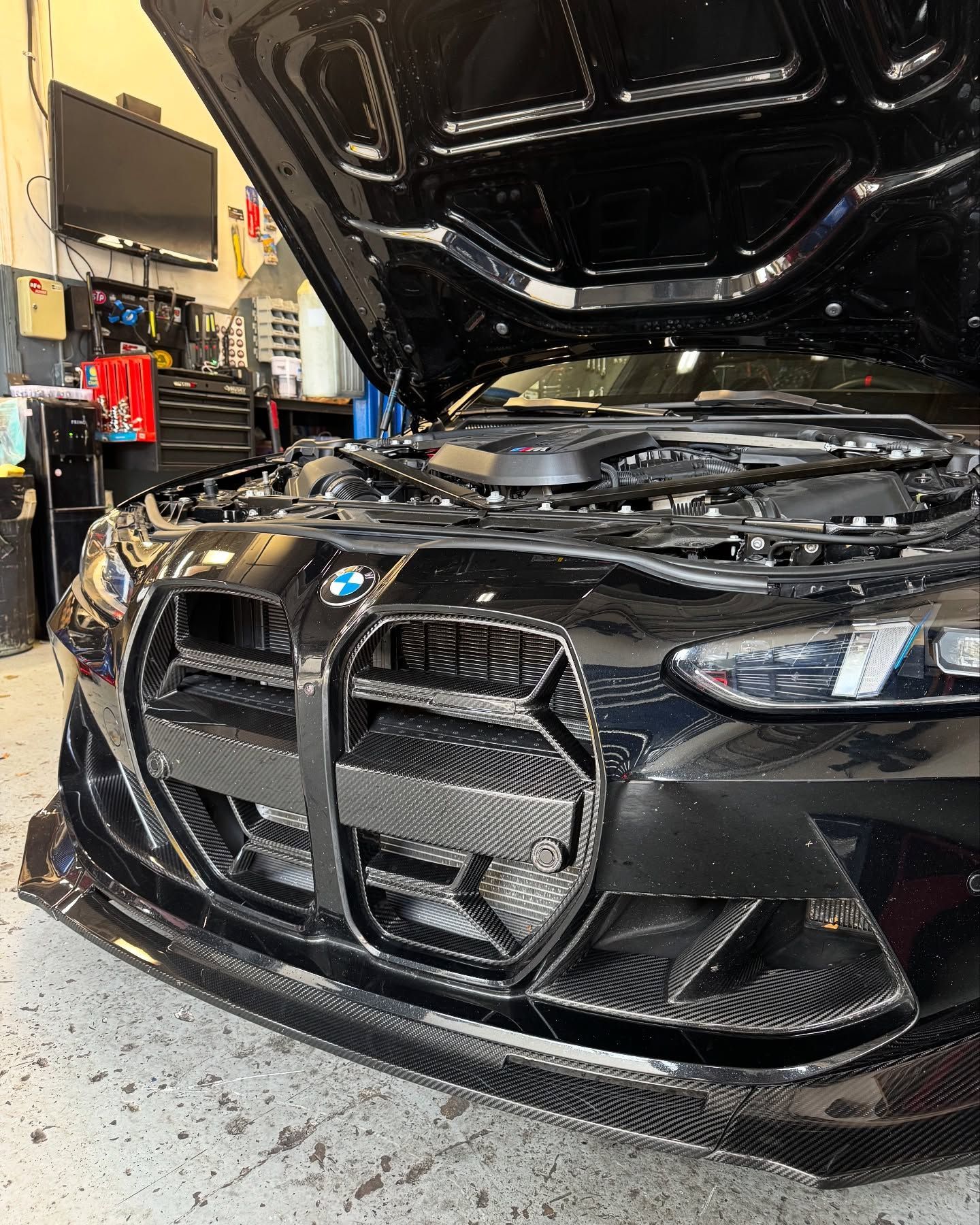 Black BMW M3 with hood open, showcasing engine bay in a garage. Carbon fiber accents visible.