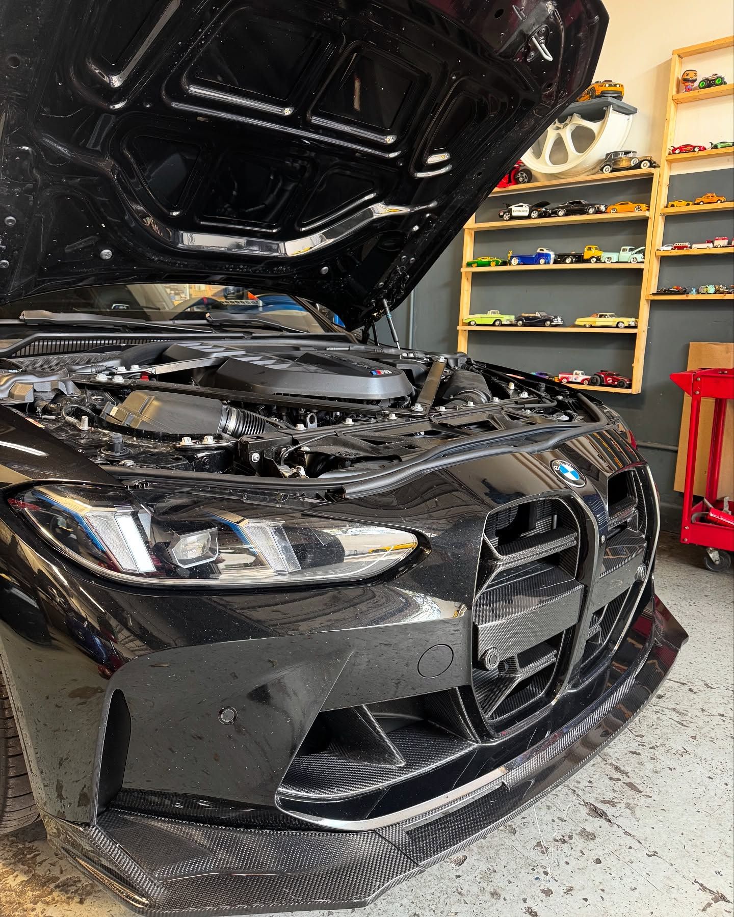 Black BMW with open hood, carbon fiber front lip, in a garage with shelves of toy cars.