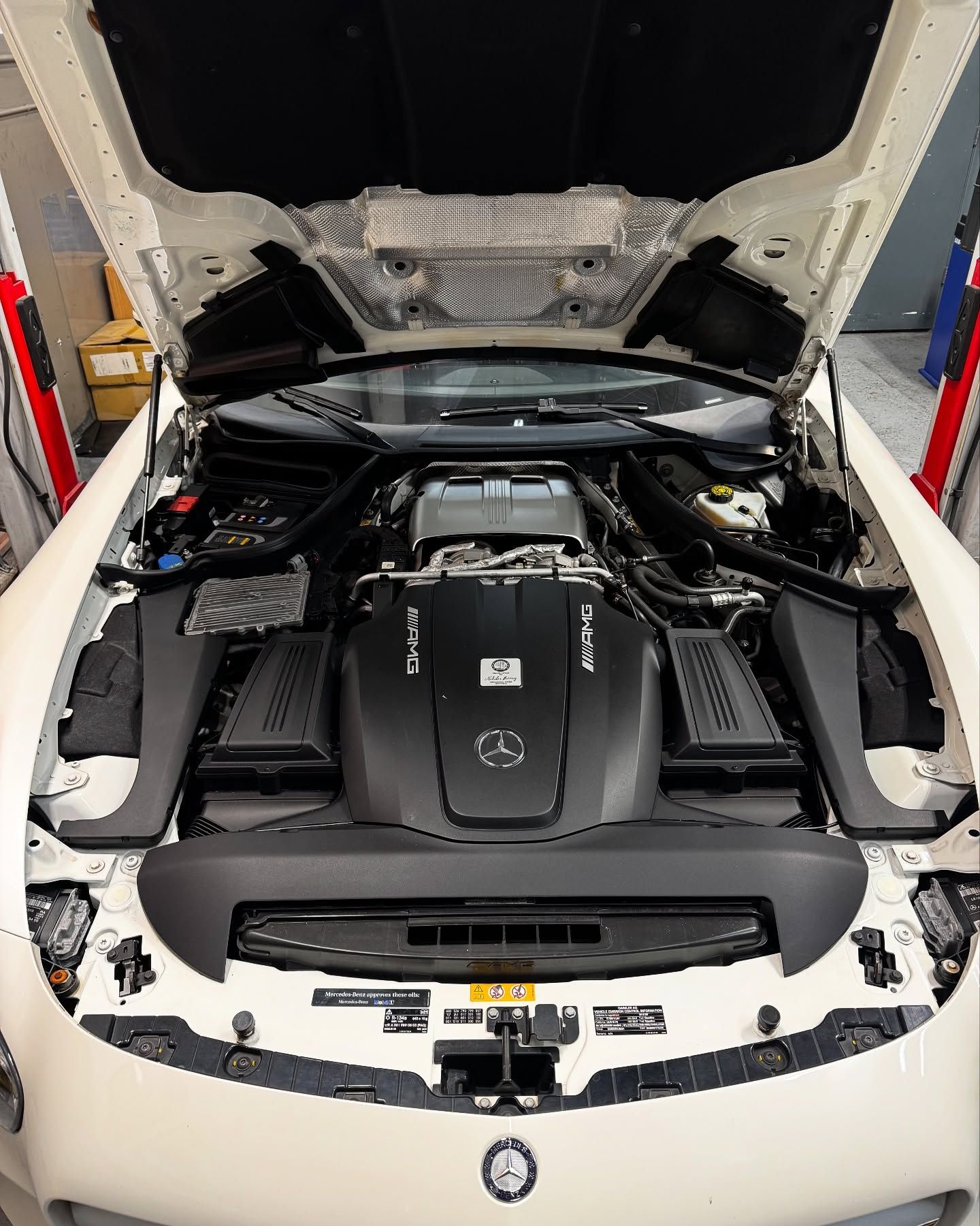 White Mercedes-AMG GT engine bay open. Dark engine cover, black side components, white exterior, in repair shop.