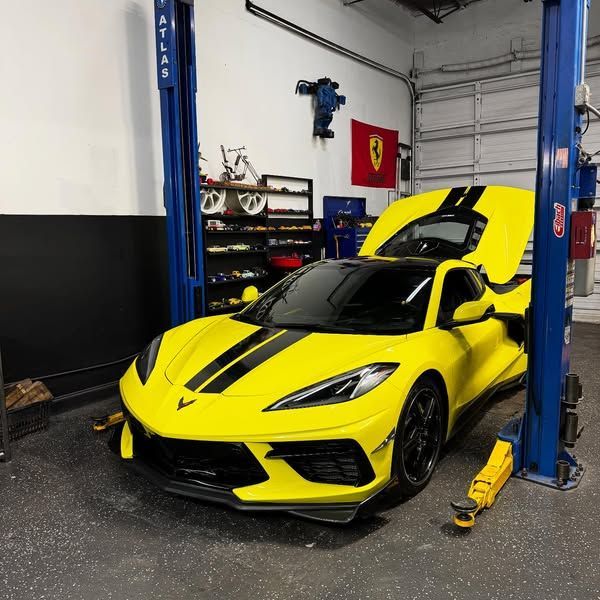 Yellow Corvette sports car on a lift in a garage. Black stripes, open hood. Ferrari emblem on wall.