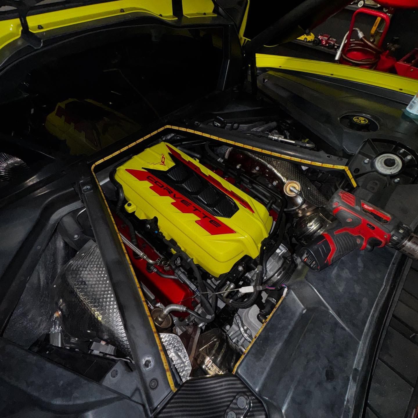 Black BMW M4 with hood open in a garage, showing carbon fiber trim, grilles, and front spoiler.