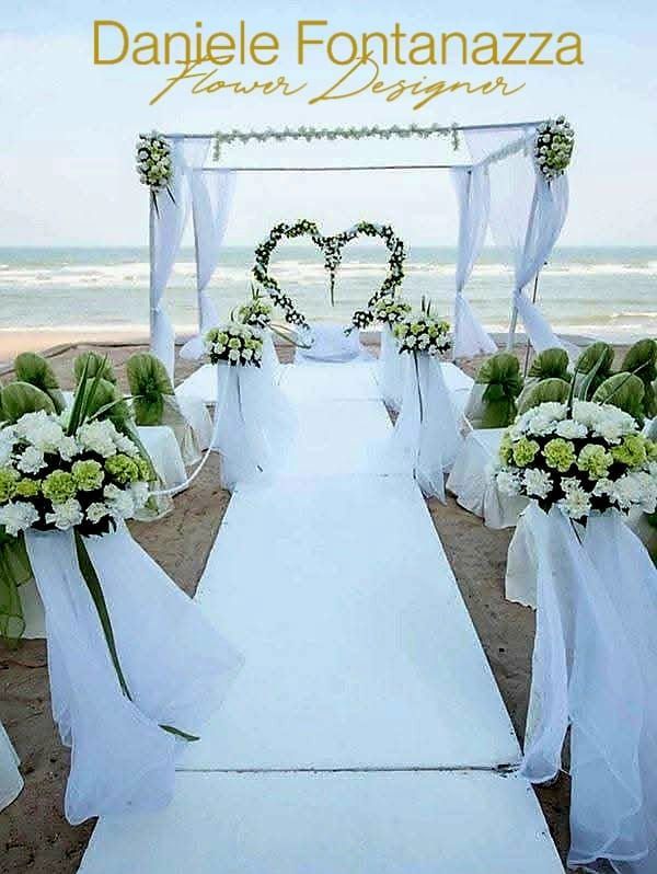 Matrimonio in spiaggia con decorazioni floreali bianche e verdi, sfondo a forma di cuore e baldacchino drappeggiato.