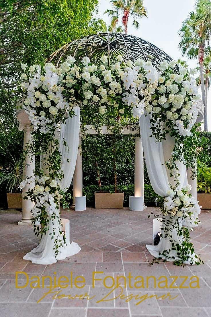 Arco nuziale floreale bianco con drappeggi e verde, all'aperto.