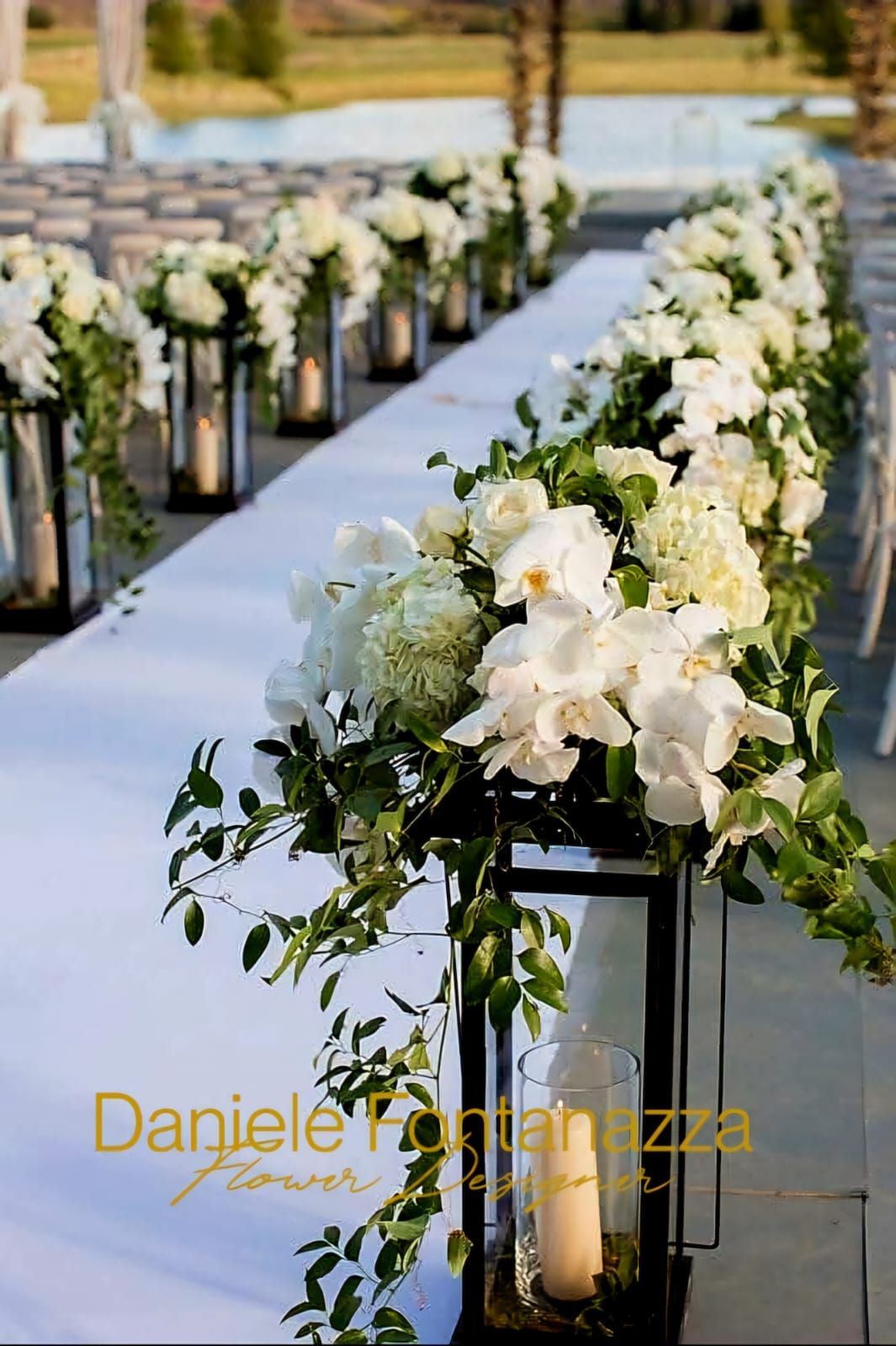 Composizioni floreali bianche in lanterne nere fiancheggiano una navata bianca, per un matrimonio all'aperto.