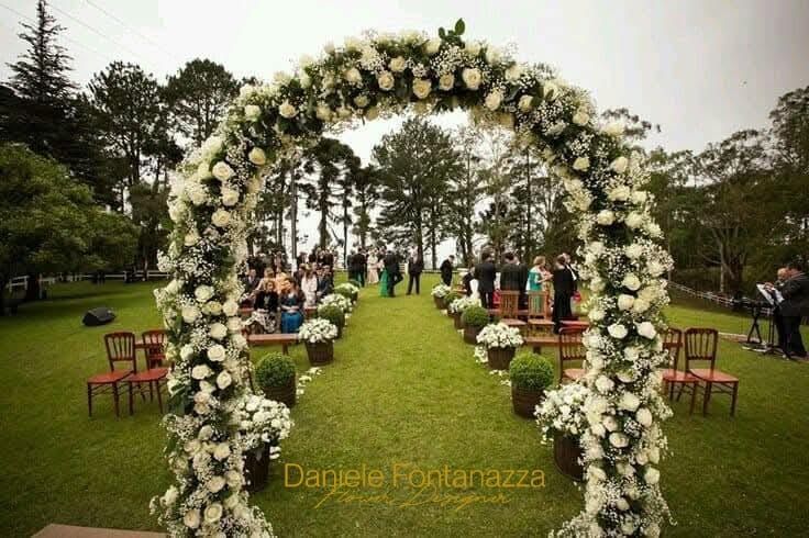 Arco per la cerimonia nuziale ricoperto di fiori bianchi su un prato verde con gli ospiti seduti sullo sfondo.