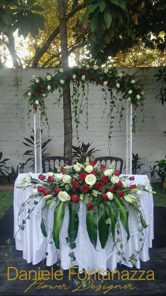 Tavolo nuziale con tovaglia bianca, centrotavola floreale e arco decorato con fiori rossi e bianchi e verde.