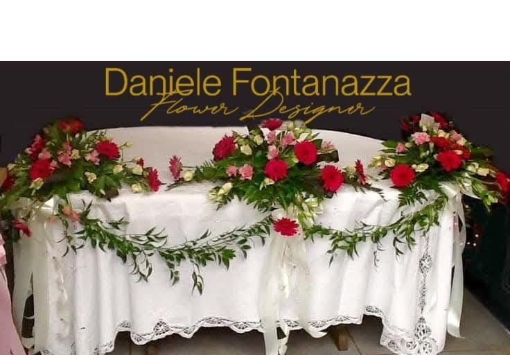 Tavolo apparecchiato per un evento, drappeggiato con un telo di pizzo bianco e decorato con composizioni floreali rosse e rosa e tralci verdi.