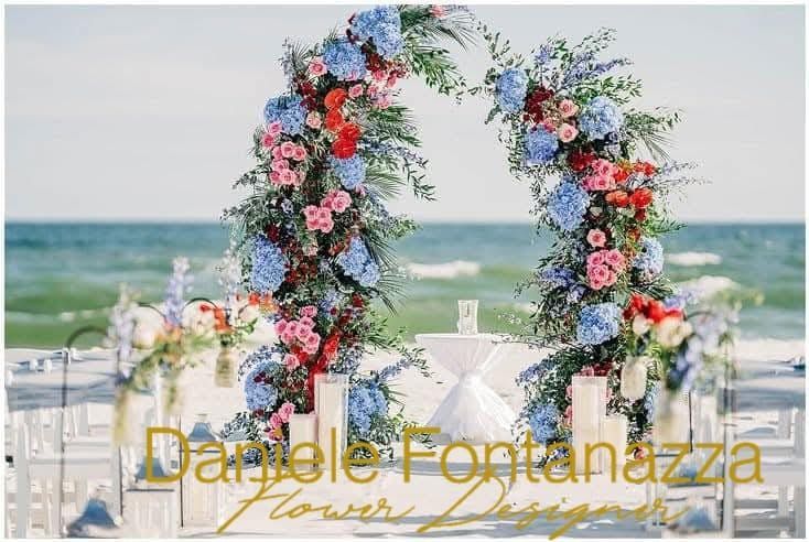 Arco floreale su una spiaggia, fiori blu e rosa, sedie bianche, oceano sullo sfondo.