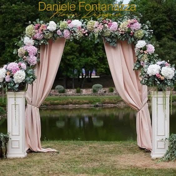 Arco floreale con drappeggi rosa, colonne bianche e lago sullo sfondo.