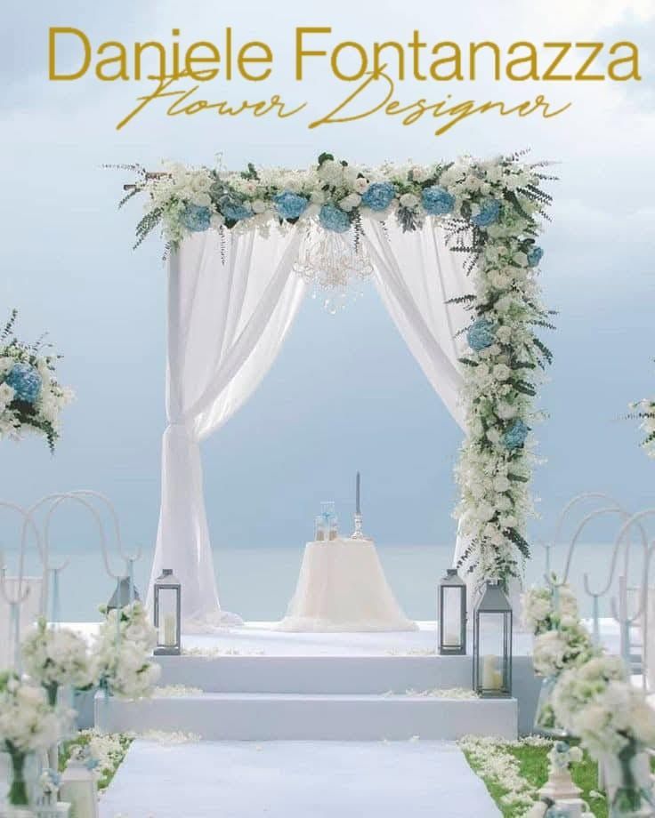 Arco nuziale decorato con fiori bianchi e blu, su una spiaggia con l'oceano sullo sfondo.