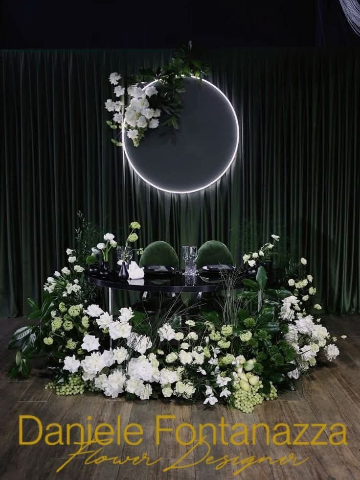 Tavolo per ricevimento di nozze con composizione floreale bianca, sfondo verde e una luce al neon circolare.