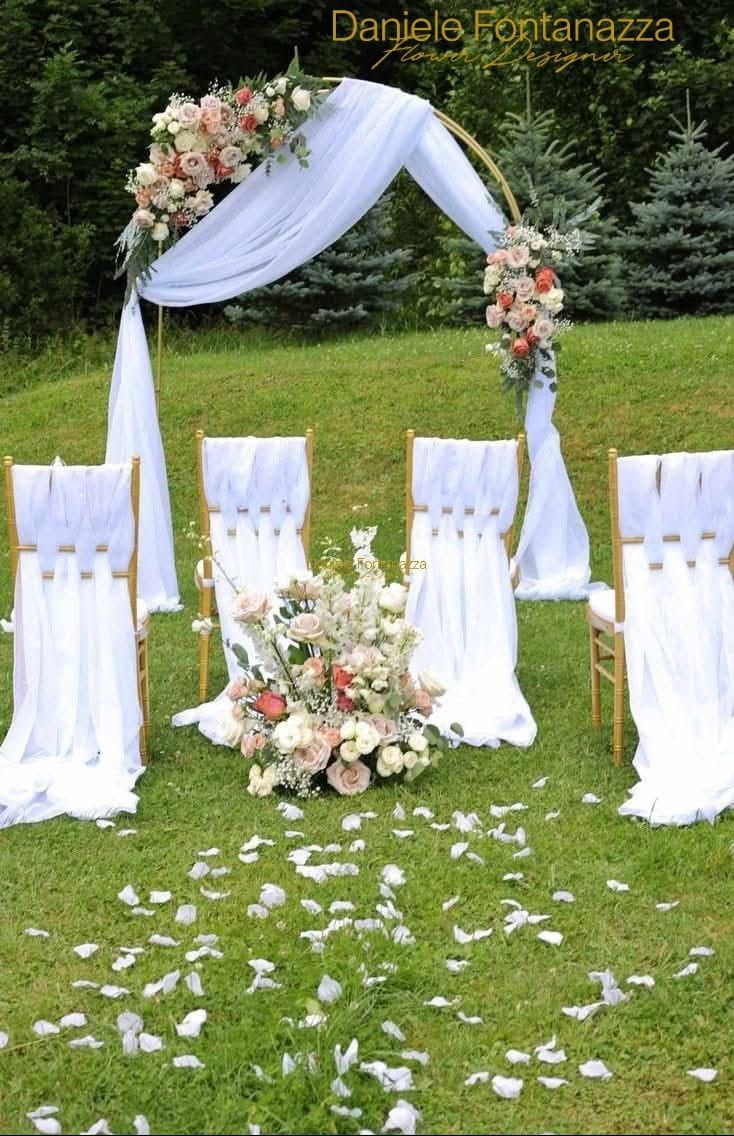 Cerimonia nuziale allestita all'aperto con arco floreale, tessuto drappeggiato, sedie decorate e petali sparsi.