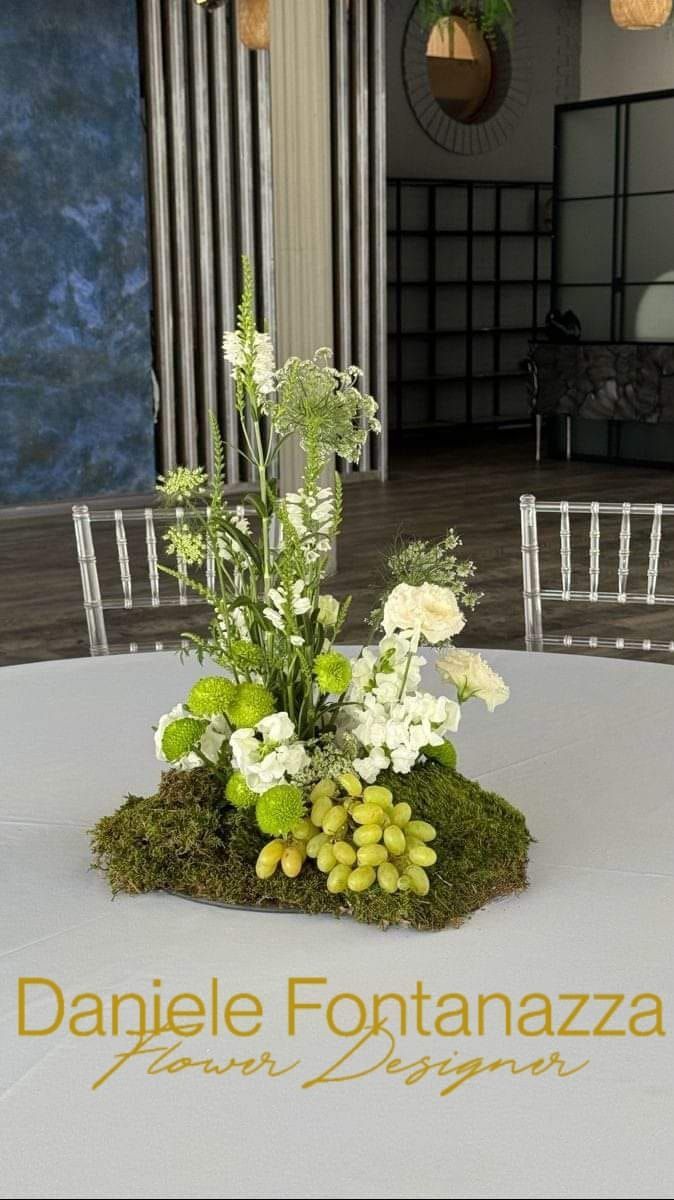 Composizione floreale su un tavolo: base di muschio, fiori bianchi e verdi, con sovrapposizione di testo dorato.
