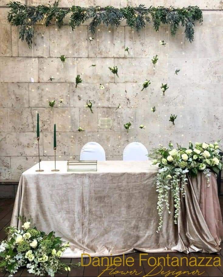 Tavolo nuziale con decorazioni floreali su uno sfondo di muro in pietra, decorato con fiori e tessuto drappeggiato.