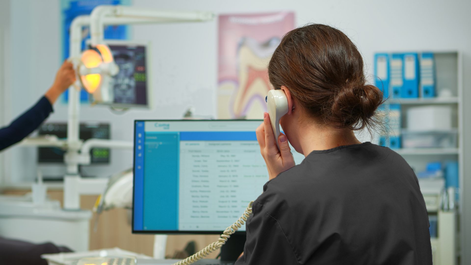 Dental office employee at computer on phone; dental light and X-ray image in background.