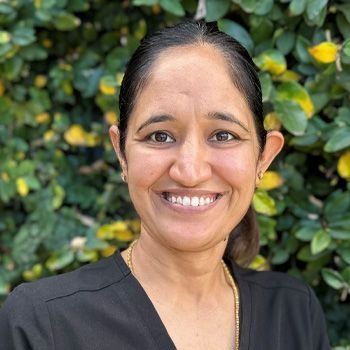 Woman with dark hair smiling in front of green foliage.