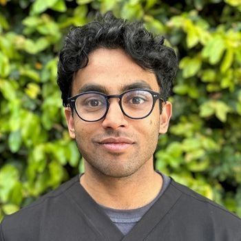 Man with dark curly hair and glasses smiles, in front of greenery, wearing a black top.