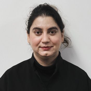 Woman with dark hair pulled up, wearing a black turtleneck and top, against a white wall.