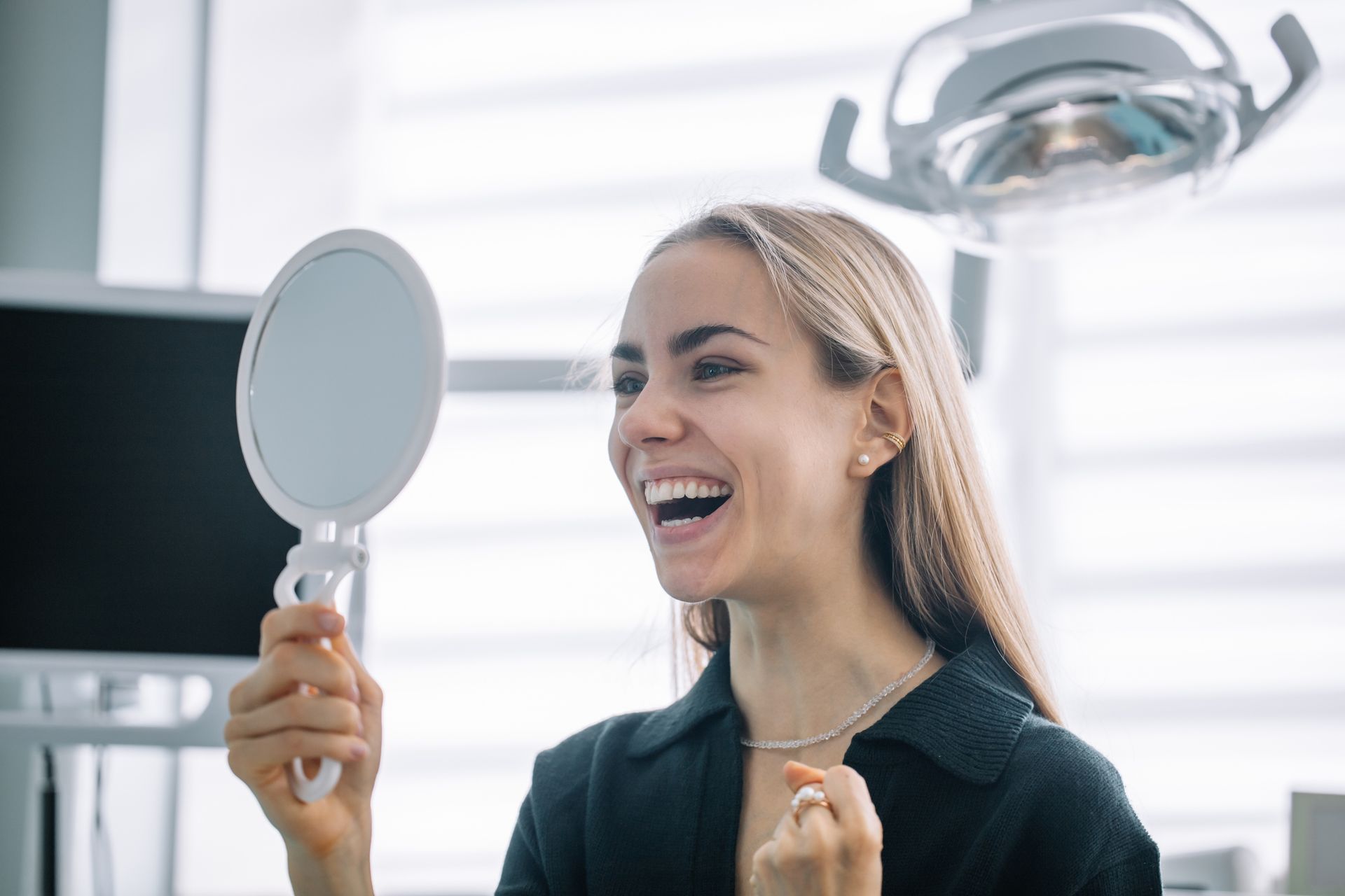 Woman smiles, holding a mirror, examining her teeth in a dental clinic.