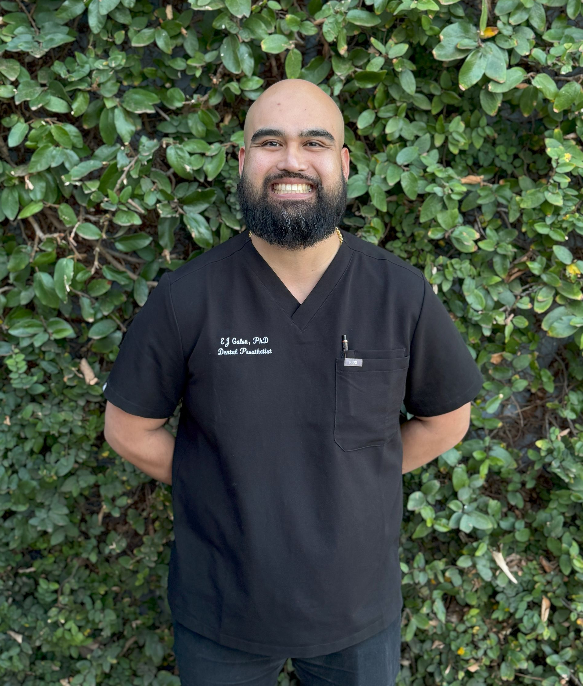 Woman in black scrubs smiling in front of green foliage.