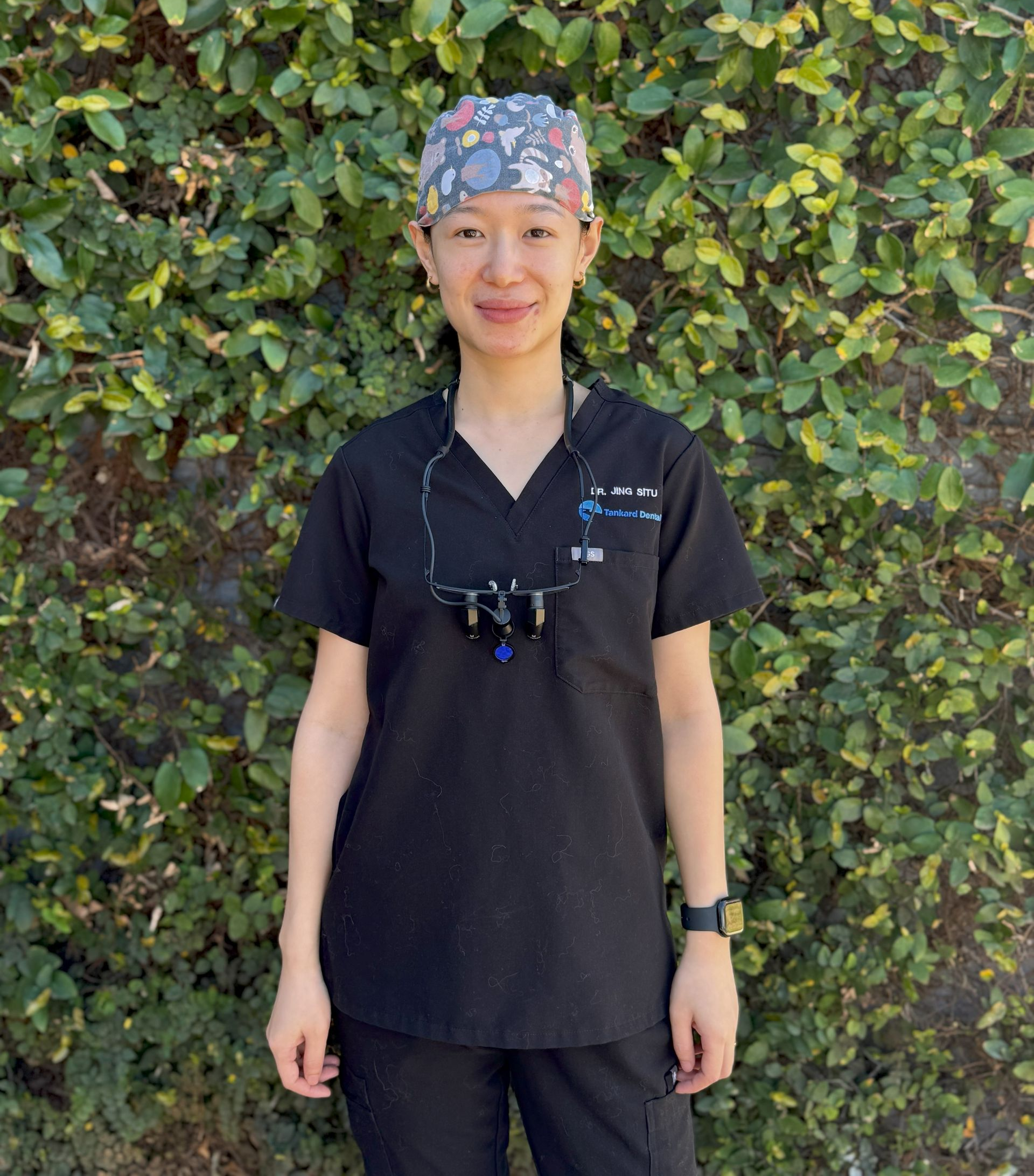 Woman in black scrubs smiling in front of green foliage.