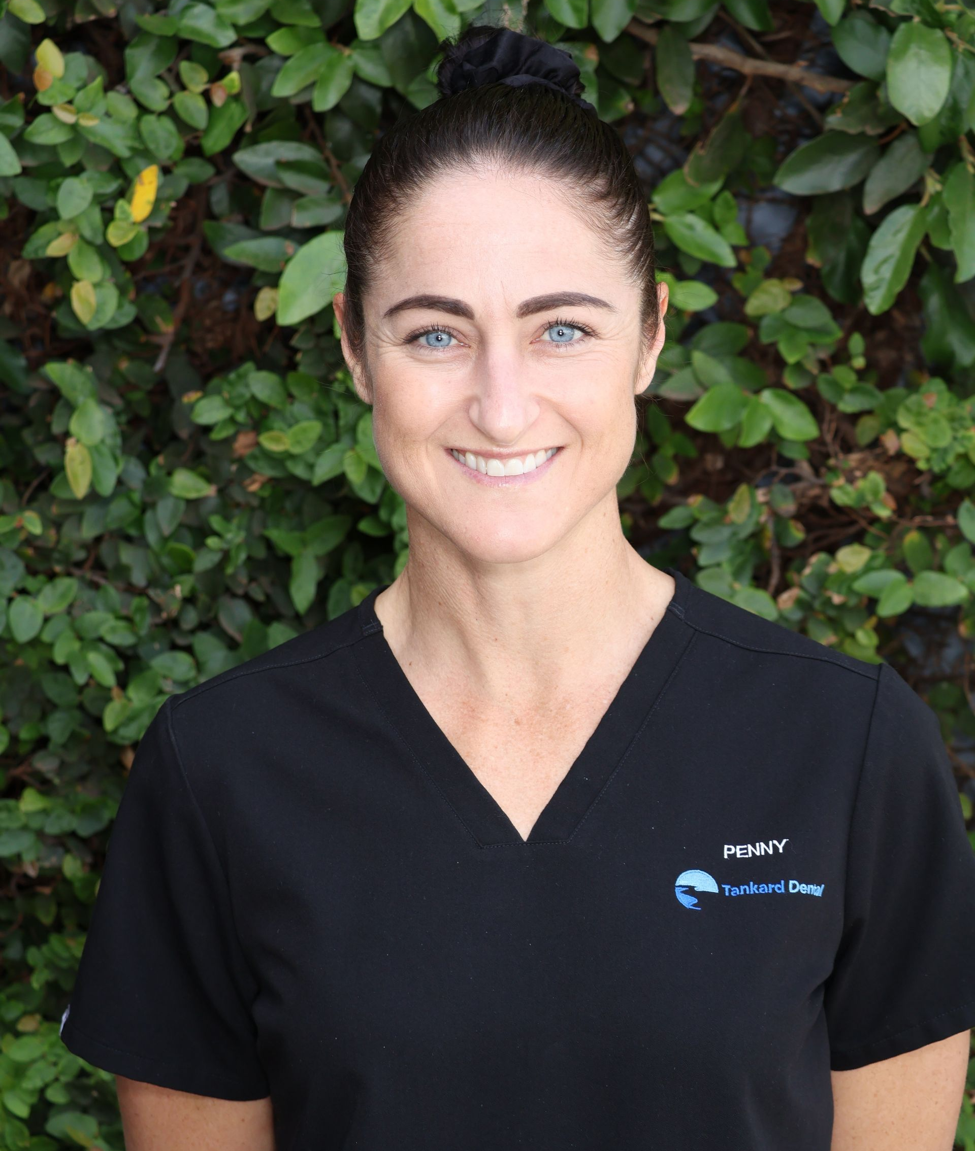 Woman in black scrubs smiling in front of green foliage.