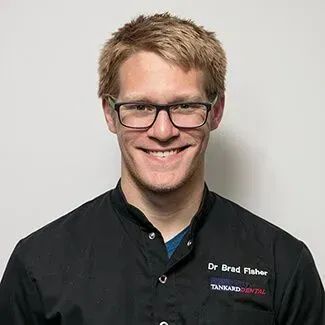 Dr. Brad Fisher smiling, wearing glasses and a black dental uniform with a logo, indoors.