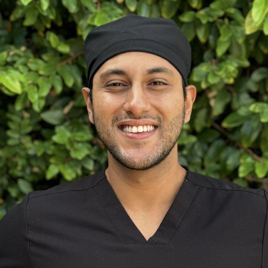 Man in black scrubs and cap smiles, against a green leafy background.