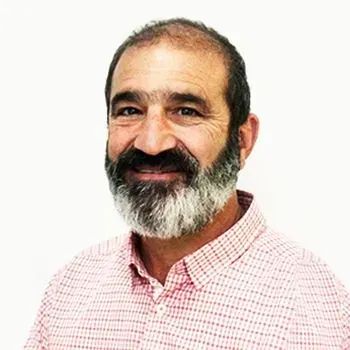 Man with a gray beard and mustache smiles, wearing a pink checked shirt, against a white background.