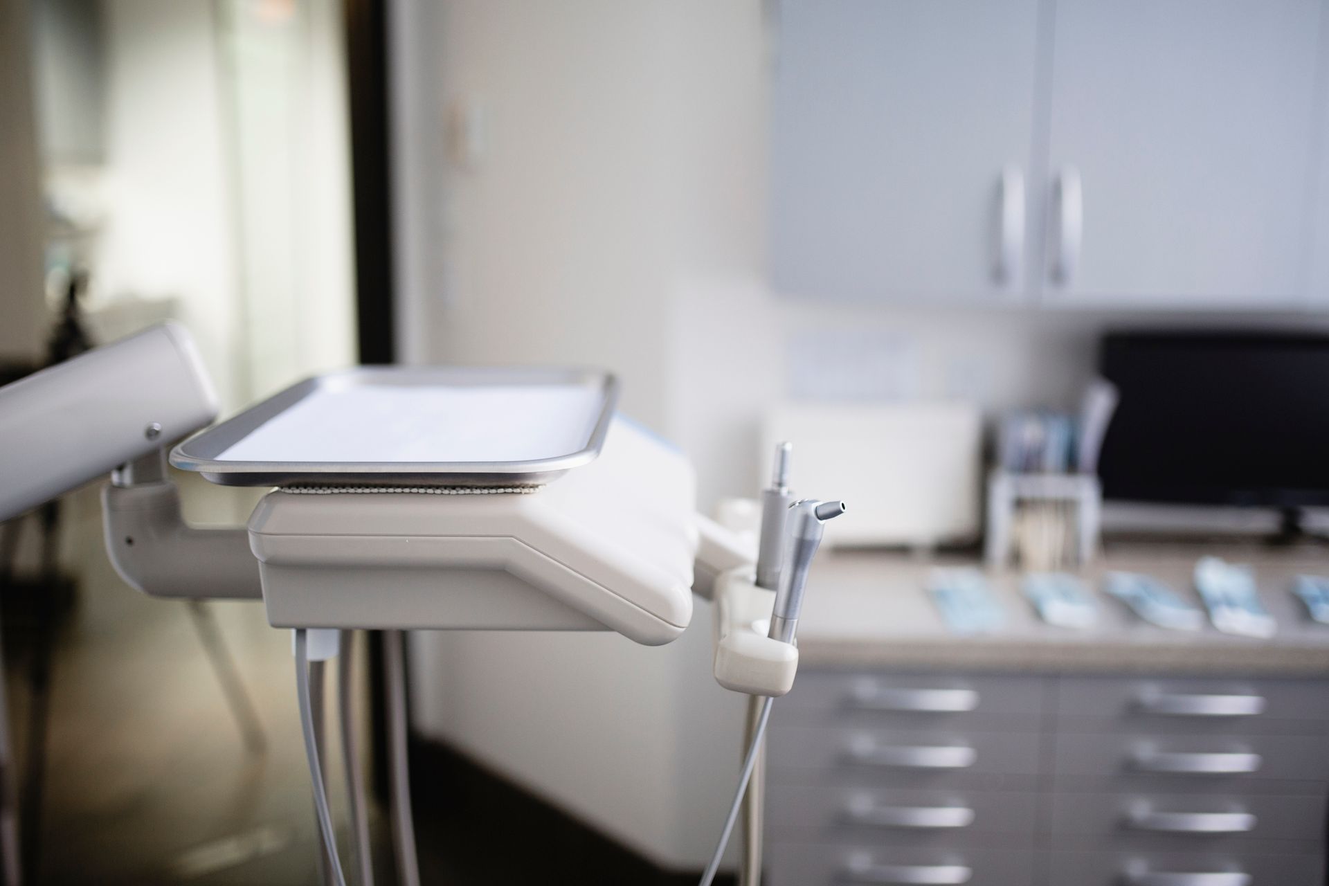Dental equipment in an examination room; stainless steel tray on a white machine; cabinets in background.
