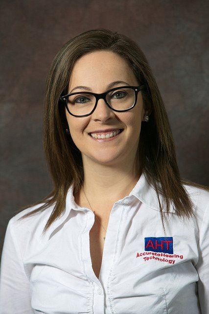 Woman with glasses smiling, wearing a white shirt with an AHT logo, against a dark background.