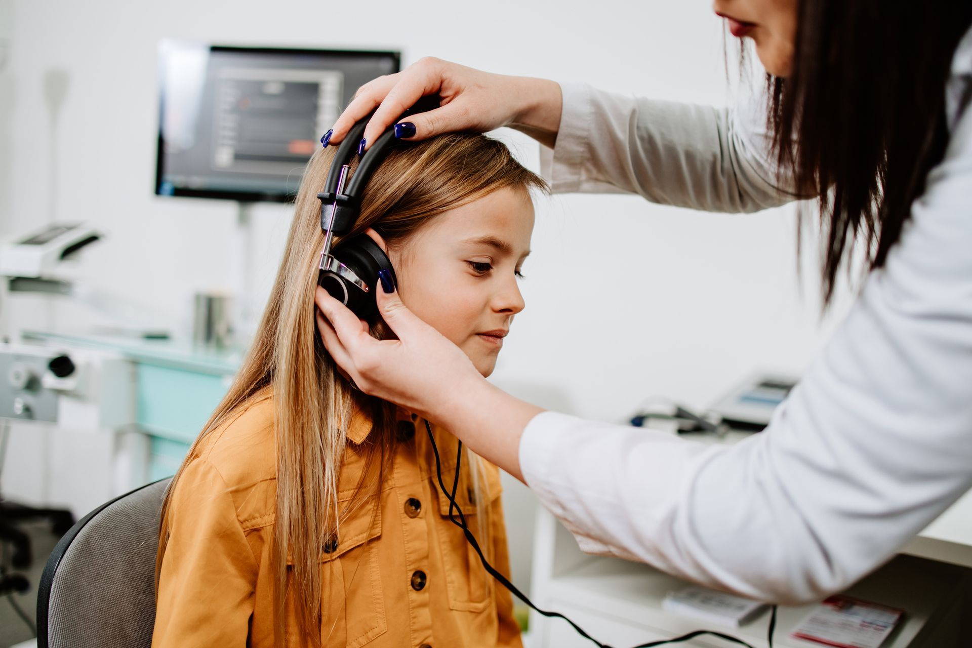 A teenage girl, in a yellow jacket, getting an auditory test at a hearing clinic from a doctor.