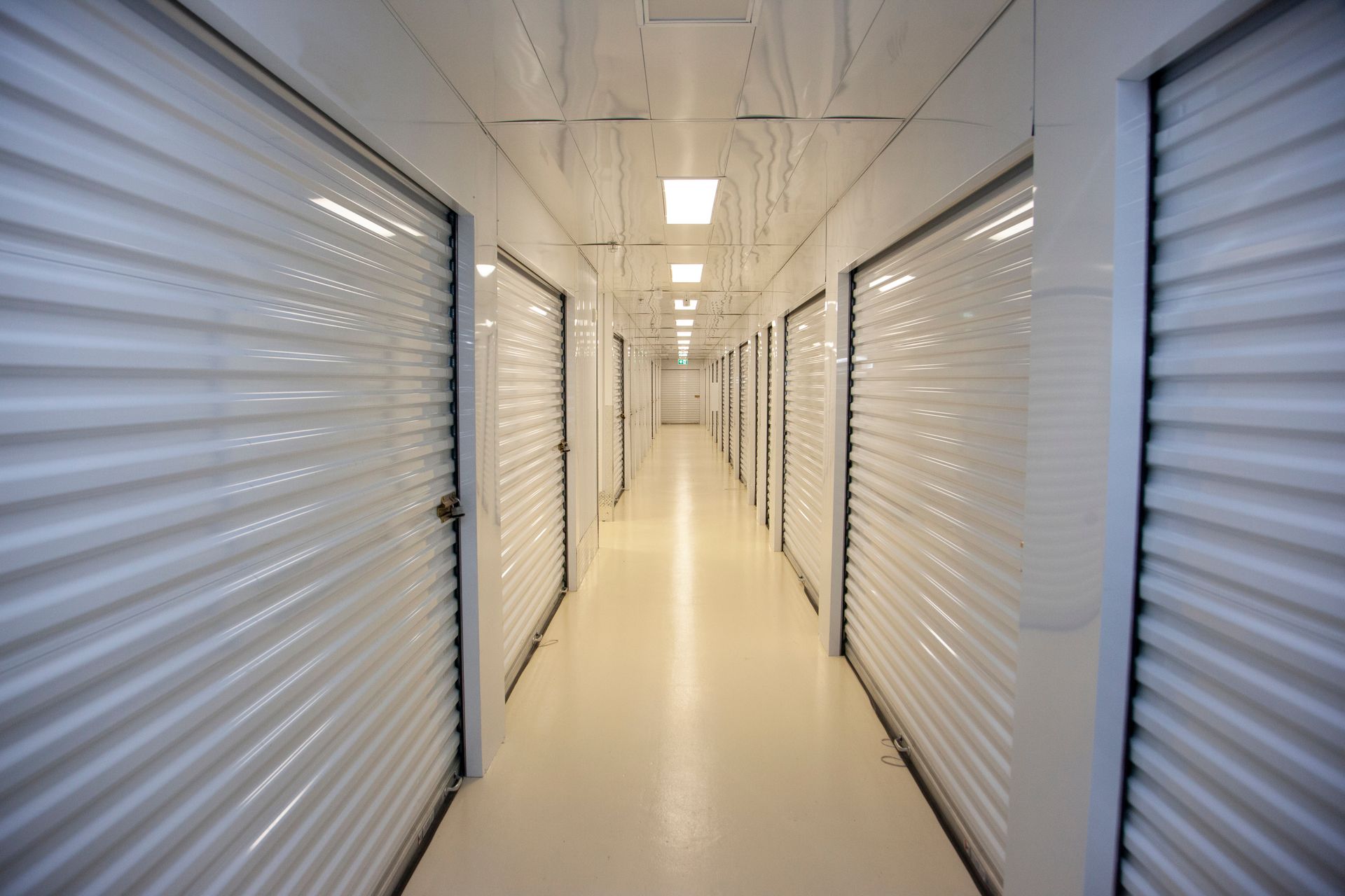 A long, narrow storage facility hallway with white roll-up doors on both sides and bright overhead lighting.