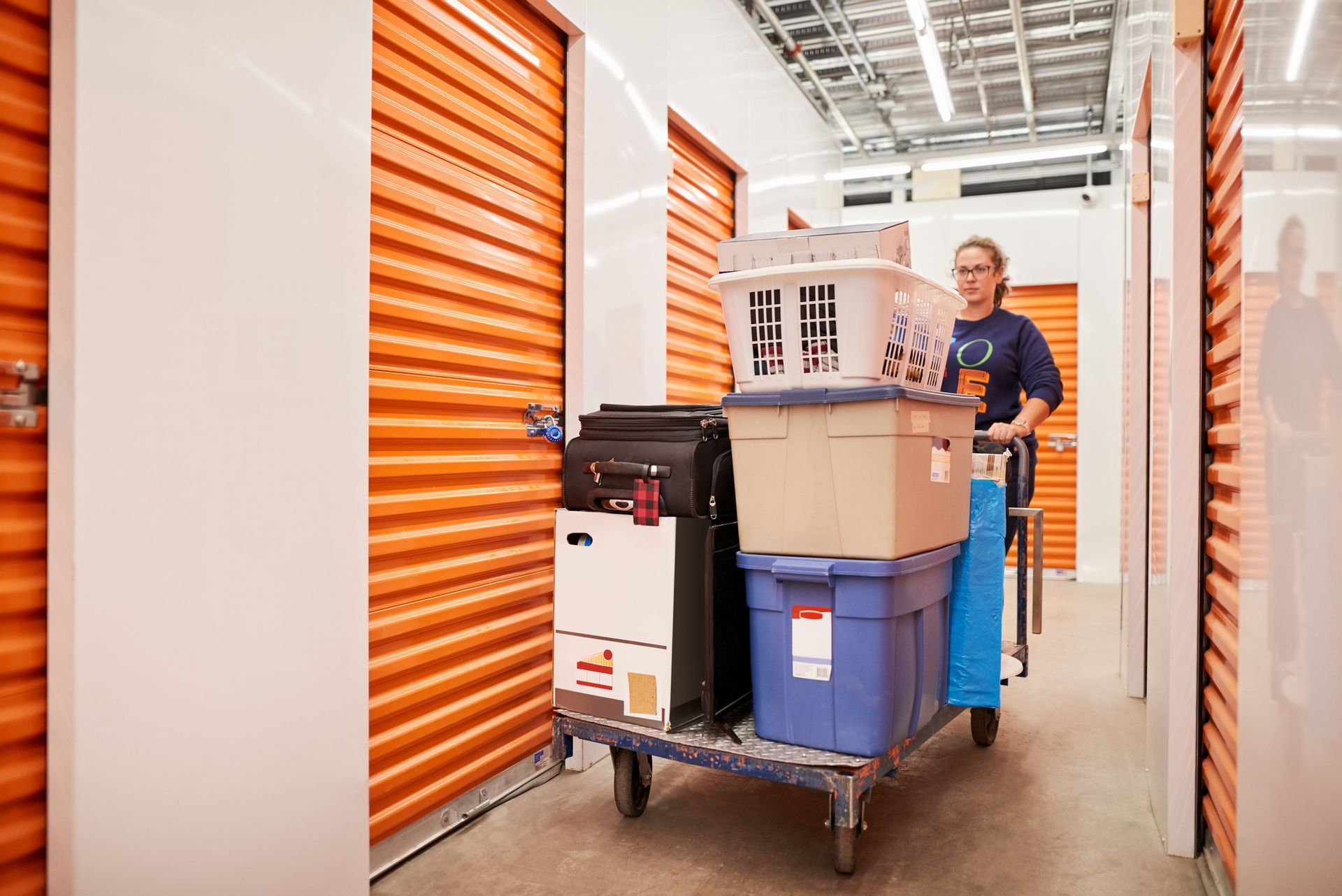 A woman is pushing a cart filled with boxes and luggage in a storage unit.