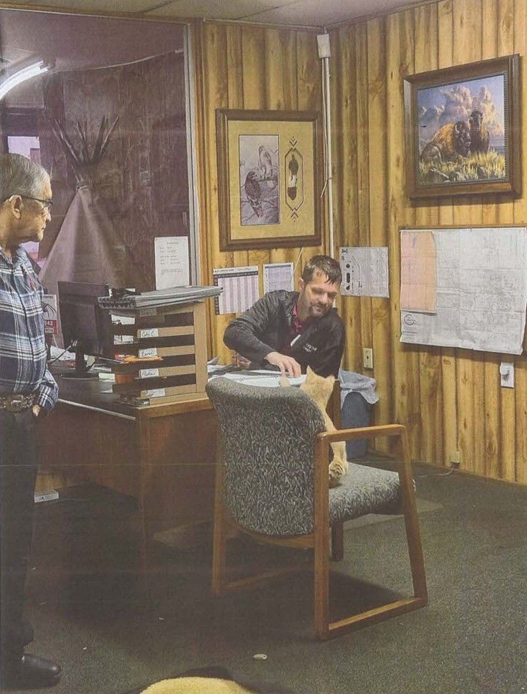 Two men in a wood-paneled office, one seated at a desk and one standing nearby with papers.