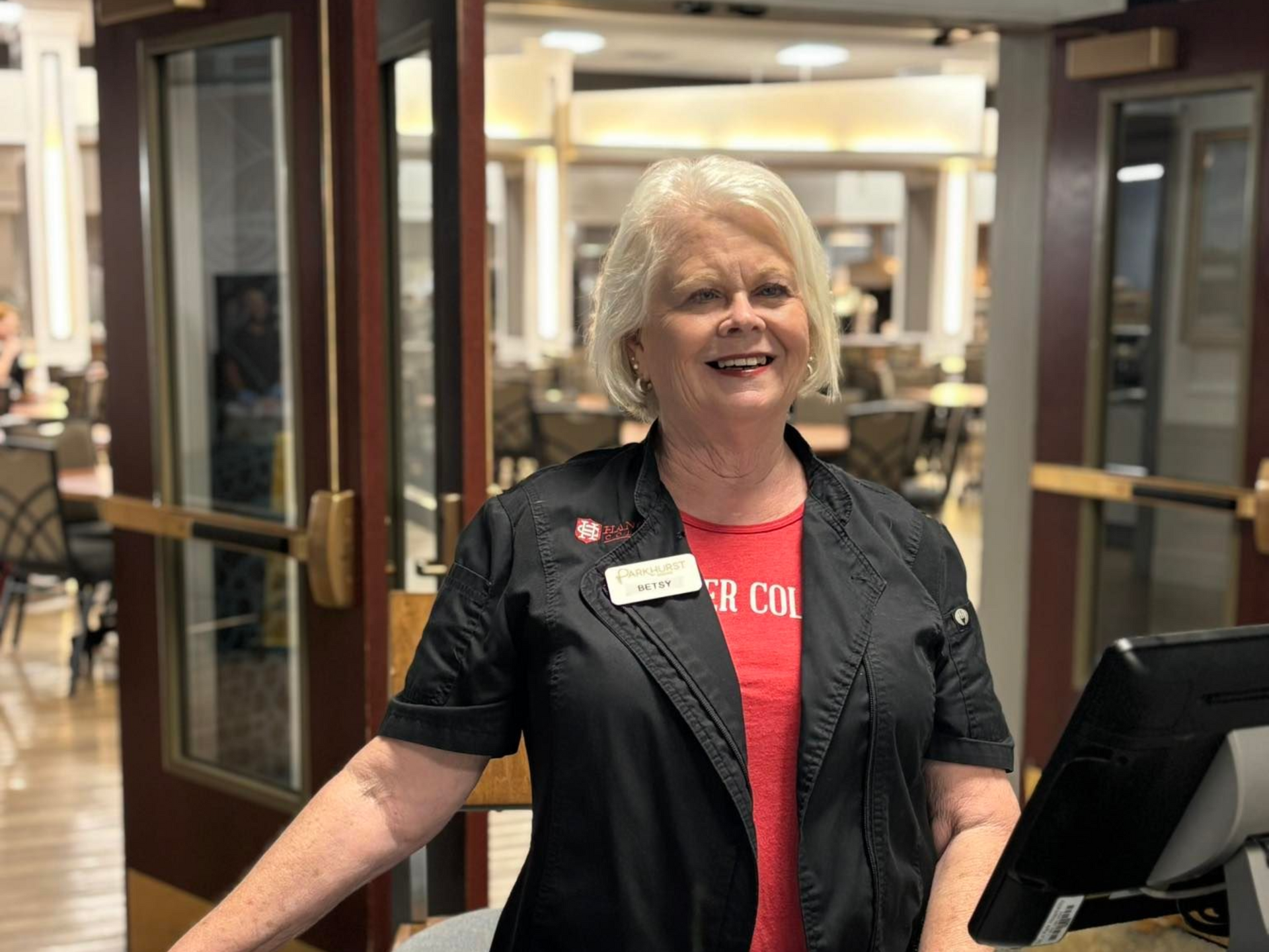 Woman in black jacket and red shirt smiling behind a counter, interior with tables.