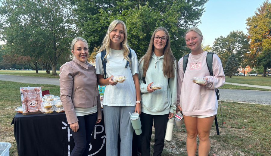 Three chefs smiling in a commercial kitchen; all wearing caps, black aprons, and blue work shirts.