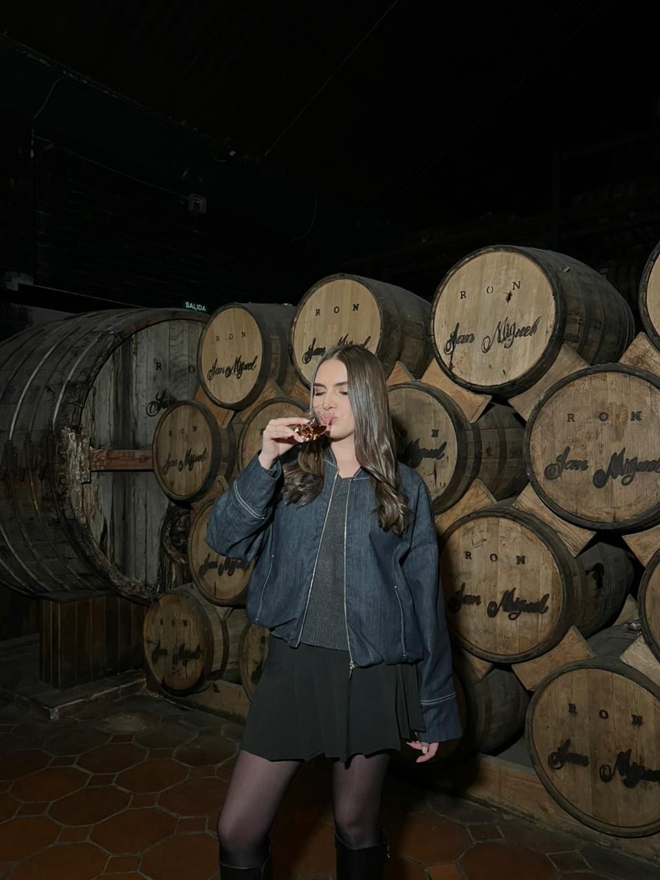 Woman in a denim jacket posing in a dim wine cellar with stacked wooden barrels.