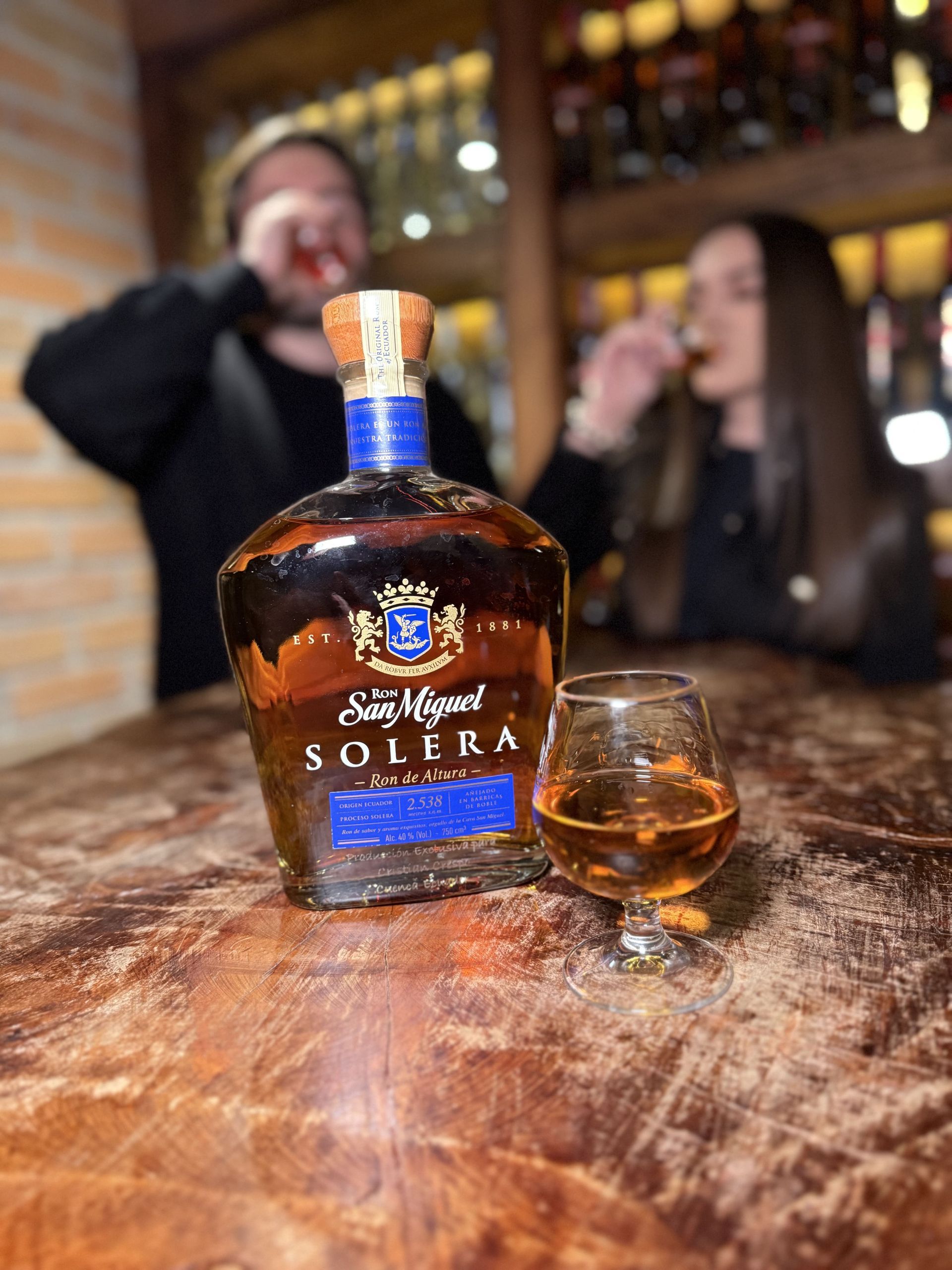 Bottle of Ron Solera rum with a filled glass on a wooden bar, with two people taking sips in the background.