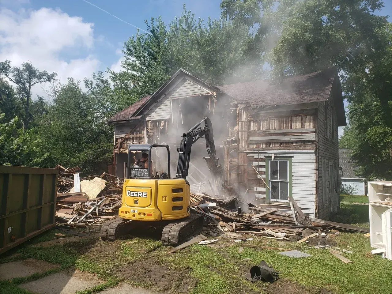 A yellow excavator is demolishing a house in a backyard.
