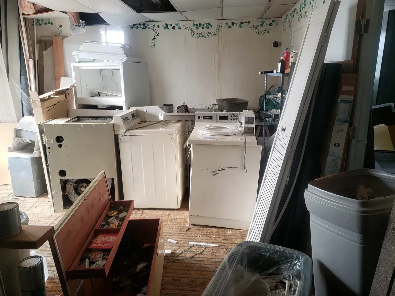 A messy room with a washer and dryer and a trash can