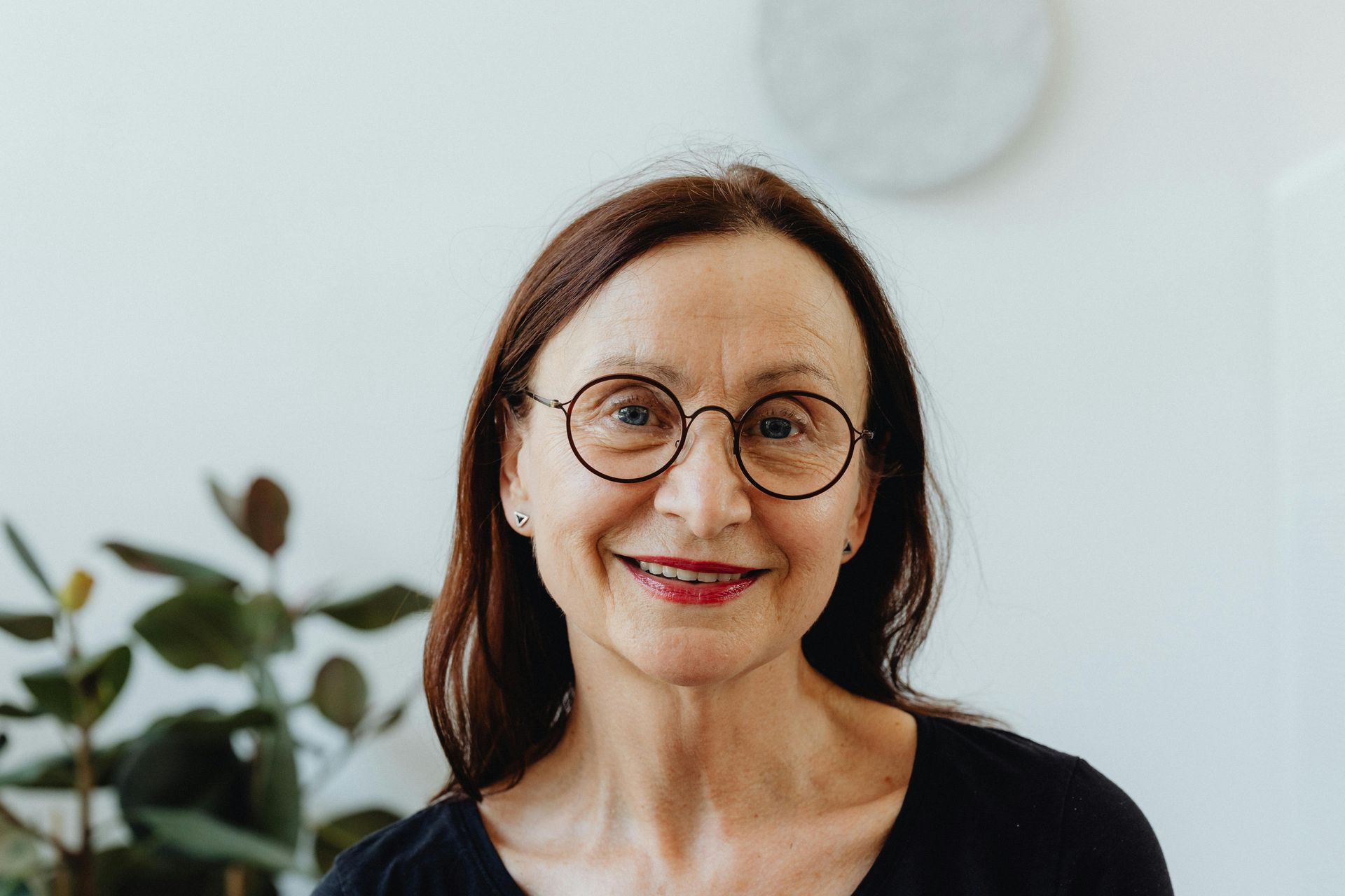 Woman with glasses smiles at the camera, in front of a white wall, with a plant.