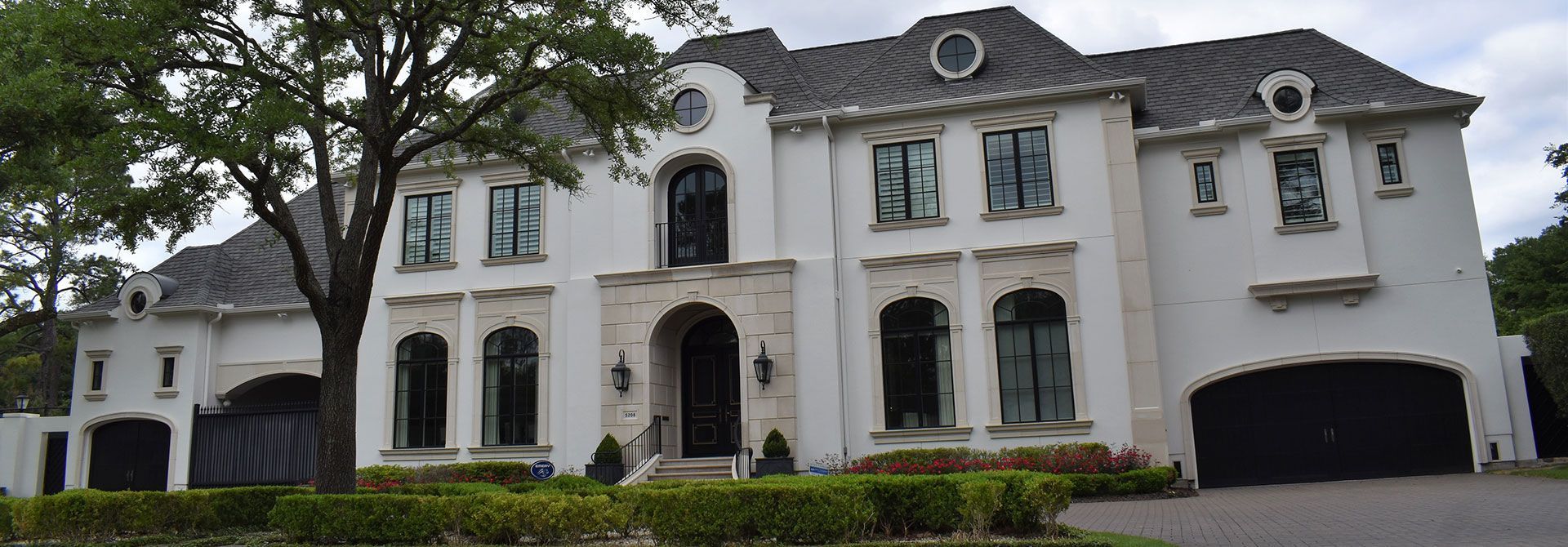 A large, white stucco mansion with a dark gray roof and black garage doors, green bushes in front.