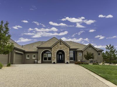Tan house with stone facade, arched entrance, and long driveway against a blue sky.