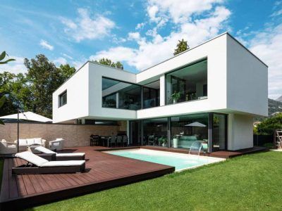 Modern white house with a pool, wooden deck, and lawn on a sunny day.