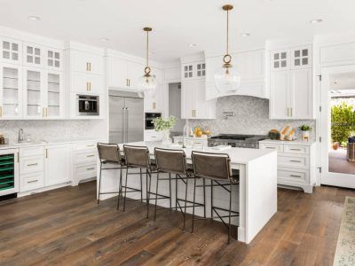 White kitchen with wooden floor, island with bar stools, stainless steel appliances, and pendant lights.