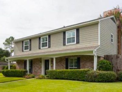 Two-story beige house with black shutters, brick facade, covered porch, and green lawn.