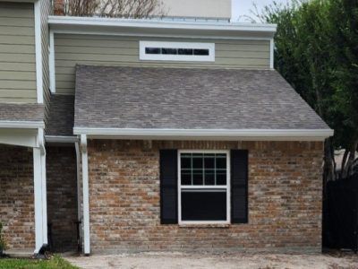 Brick building with a dark shingled roof, window with black shutters, and light green siding.