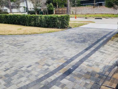 Brick paver driveway with dark border, next to green hedge and lawn.