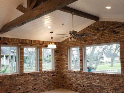 Brick-walled sunroom with white ceiling, wood beams, and multiple windows overlooking a yard.